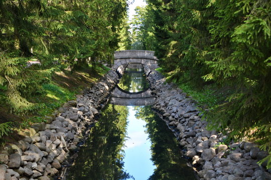 Old round stone bridge in the coniferous forest over a long narrow ditch with blue water between the stone walls in a beautiful city garden. Horizontal frame
