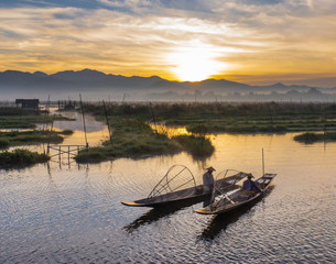 two Intha fishermen warming themselves by the fire on their boats during early morning sunrise on Lake Inle, Myanmar, Burma