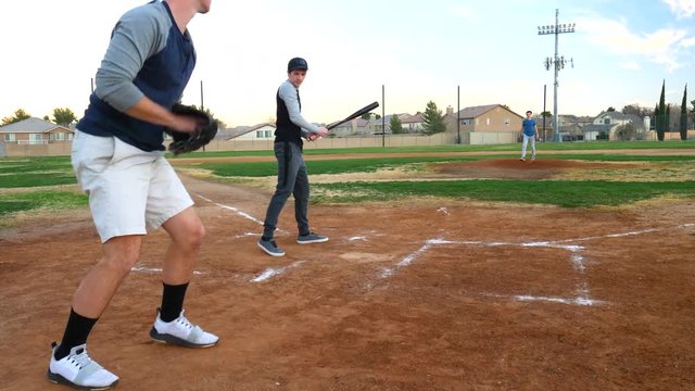 A pitcher throwing a strike to a young man at bat at home plate during an amateur baseball game at sunset in a park field.