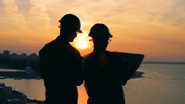 Workers talk, standing on a sunset background, back view.