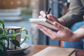 Closeup image of a woman's hands holding and writing on blank notebook with coffee cup on wooden table