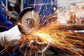 Close-up on the sides fly bright sparks from the angle grinder machine. A young male welder in a...