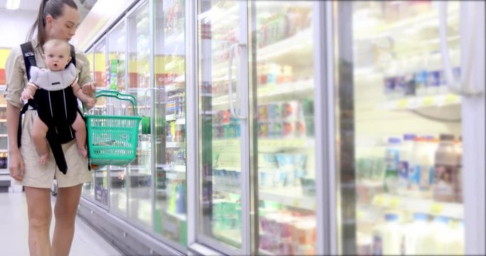 Mother with child in ergo backpack chooses dairy products from refrigerator in store