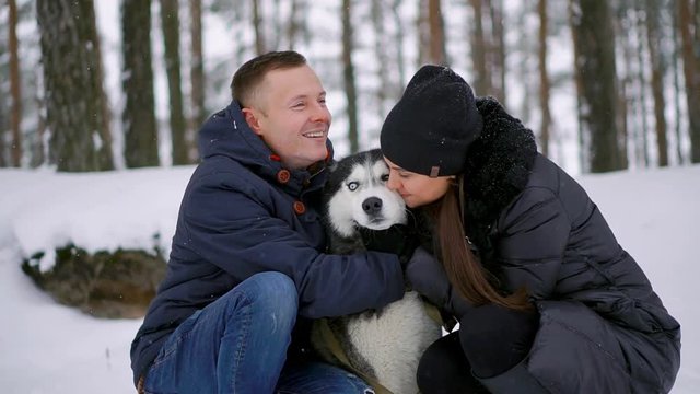 Family portrait of cute happy couple hugging with their alaskan malamute dog licking man's face. Funny puppy wearing santa christmas deer antlers and kissing woman. Freedom lifestyle pet lovers.