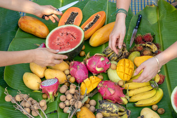 Tropical fruits assortment on a green banana leaves and people hands. Yummy dessert, close up. Mango, papaya, pitahaya and hands, top view