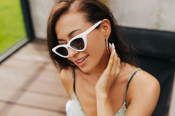 Fototapeta premium Close-up portrait of beautiful tanned girl in trendy sunglasses lyuing by swimming pool outside. Elegant cheerful young woman enjoying sunny, while resting outdoor on summer resort