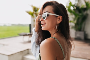 Exited happy woman with long wavy hair wearing fashion glasses and stylish earrings on background of resort and pool