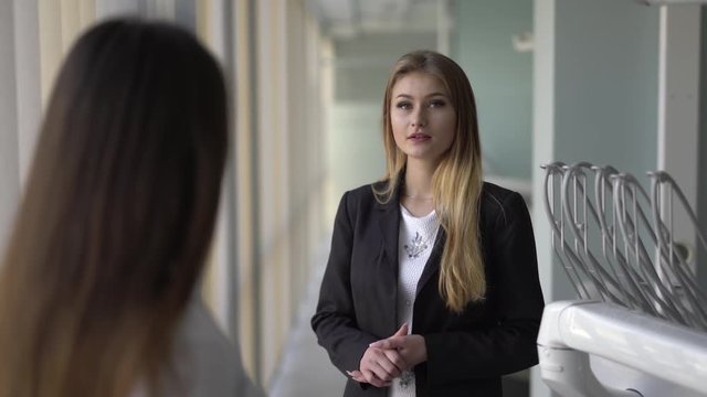 Nurse standing with visible back and the female client explaining the doctor her problem, poining to her right/left cheeck where it being hurt/toothpain. Blond girl well-dressed and slim