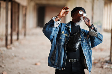 Fototapeta na wymiar African american man in jeans jacket, beret and eyeglasses, smoking cigar at abandoned factory.