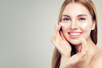 Exited beautiful woman with healthy clear skin applying moisturizing cream on her face, closeup...