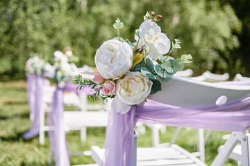 Close up of floral composition on white wooden chairs with violet cloth on each side of archway outdoors, copy space. Empty chairs for guests prepared for wedding ceremony