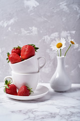 Ripe organic strawberries in white ceramic bowl and chamomile flowers in vase on gray concrete background, copy space. Healthy food concept, still life