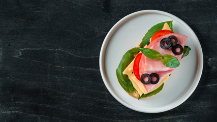 Fresh sandwich with ham, cheese, olives and basil leaves on white plate ready-to-eat. Slice crust baguette bread with perfect ham on black background. Copy space for text. Top view or flat lay