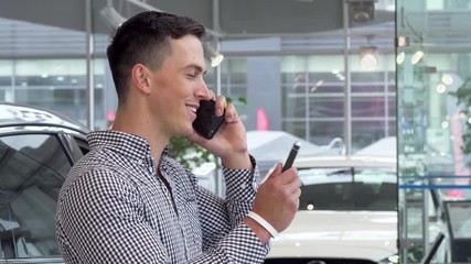 Excited man smiling, holding car keys at the dealership, talking on the phone. Handsome male driver celebrating buying new automobile, calling to share news, using smart phone