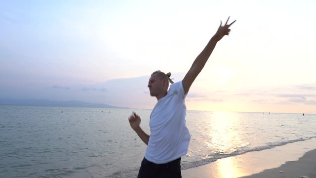 Young guy with dreadlocks dancing during sunset on the tropical beach near sea water. Happy handsome man with dreadlocks his head, slow motion