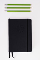 Top view of black journal with three green pencils on white background © Mathieu