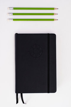 Top view of black journal with three green pencils on white background