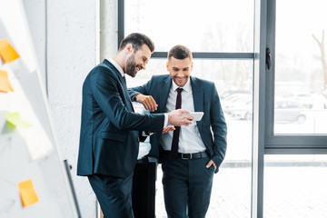 handsome businessman looking at smartphone with colleague standing with hand in pocket