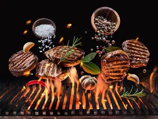 Fotobehang Gegrilde biefstukken met groenten en kruiden vliegen over het gloeiende grillbarbecuevuur. © volff