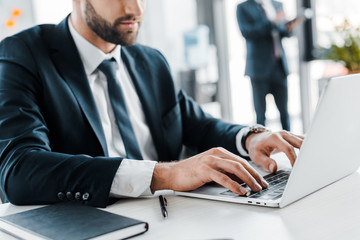 cropped view of businessman typing on laptop with coworker on background