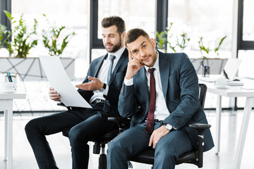 selective focus of bored man in formal wear sitting near coworker with paper in hands