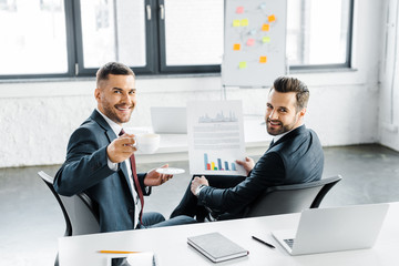 selective focus of cheerful businessman holding paper with graphs near happy coworker with cup