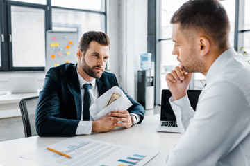 bearded businessman holding envelope with bribe near coworker in modern office