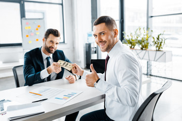 cheerful businessman showing thumb up while taking dollar banknotes in modern office