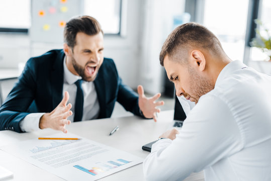 selective focus of upset man sitting near angry man in formal wear screaming in modern office