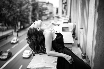 Black white portrait of young woman in elegant silk black dress lookingto a city from a balcony, fashion beauty photo
