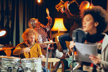 Mixed race woman singing. In background band playing instruments. Home studio interior. Selective focus on background.