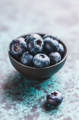 Fresh ripe blueberry in a small black bowl on a textured blue table. Macro photography.
