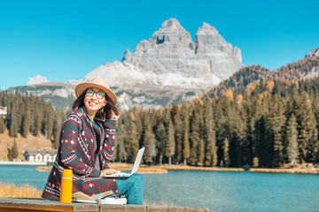 Happy Asian woman working on laptop on mountain lake background in autumn. The concept of remote...