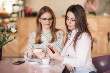 Two girls sitting in a cafe drinking cappuccino and using smartphone. Modern girl