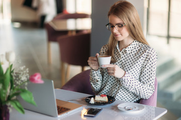 Girl eating cake in a cafe. Blonde girl in polka-dot shirt with laptop. Drinking cappuccino. Business lady
