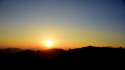 Picture of a sun setting behind a dense forest area followed by mountains.
