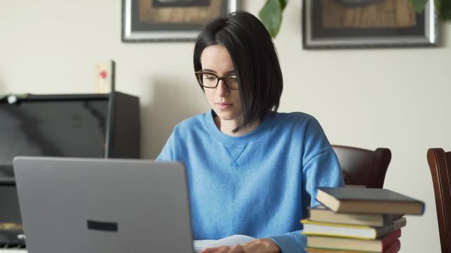 Very beautiful girl with blach hair in glasses in blue pullower typing on laptop with serious face expression. Girl starring in computer screen. Books around the table