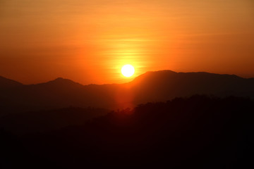 Picture of a sun setting behind a dense forest area followed by mountains.