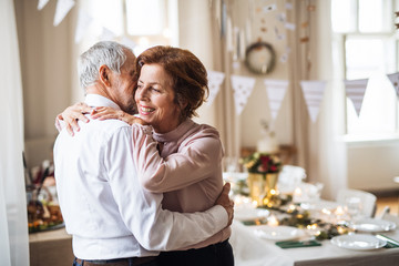 Senior couple standing indoors in a room set for a party, hugging.