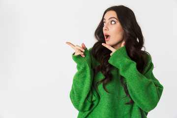 Shocked young woman dressed in green sweater posing isolated over white wall background pointing.