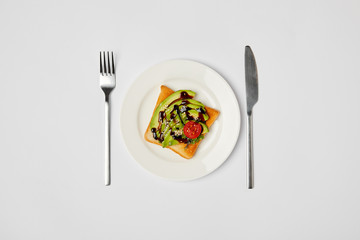 top view of toast in plate with avocado and cherry tomato, fork and knive on grey background