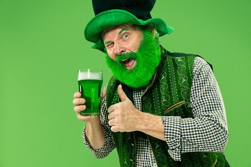 A smiling happy senior man in a leprechaun hat with green beer at studio. He celebrates St....