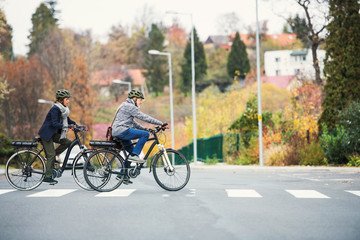Active senior couple with electrobikes outdoors crossing a road in town.