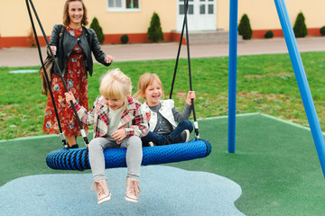 Happy family having fun together on playground. Happy childhood. Family vacation. Joyful little girls enjoying at modern swing. Young mother with daughters at park. Little girlfriends playing outdoor.