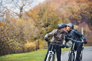 Active senior couple with electrobikes standing outdoors on a road in nature, kissing.