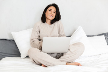Photo of mature woman 30s using laptop, while sitting in bed with white linen in cozy apartment
