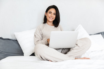 Photo of joyful woman 30s using laptop, while sitting in bed with white linen in cozy apartment