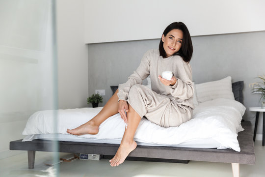 Image of attractive woman 30s sitting on bed with white clean linen in bedroom, and applying body cream on her legs