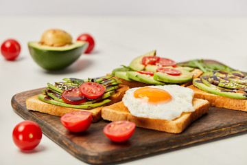 selective focus of wooden cutting board with toasts, scrambled egg, cherry tomatoes and avocado on grey background