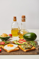 selective focus of wooden cutting board with toasts, scrambled egg, cherry tomatoes, avocados and oil bottles on grey background